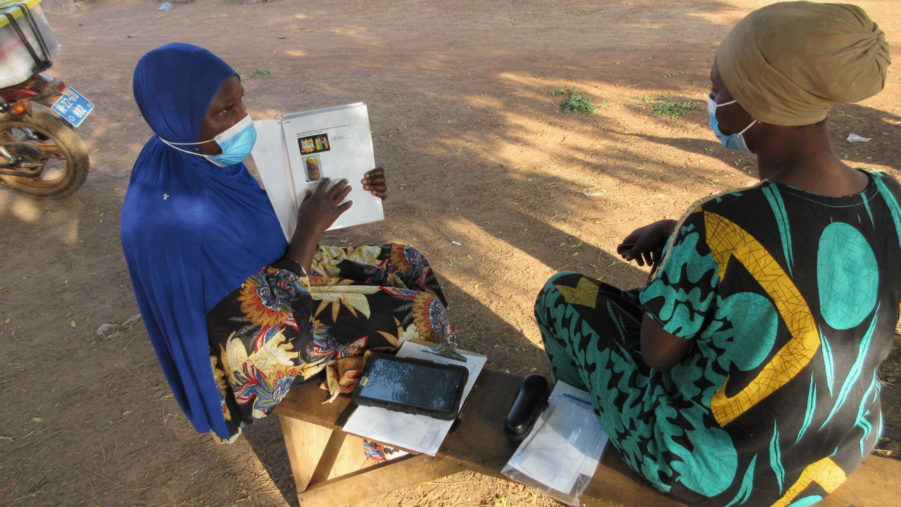 woman holding up and pointing to a document for another woman, both in traditional african style dress